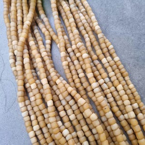 Peut inclure: Gros plan sur plusieurs brins de perles carrées brun clair. Les perles sont enfilées ensemble, créant une surface texturée. Une feuille verte est visible en haut de l'image. Les perles ont une forme et une couleur légèrement irrégulières.