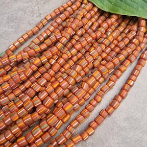 Pode incluir: Um fio de contas de vidro listradas de laranja, verde e branco. As contas são pequenas e quadradas.