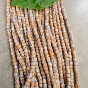 Może przedstawiać: Zbliżenie wielu pasm cylindrycznych koralików. Koraliki są głównie beżowe z pomarańczowymi i niebieskimi paskami. Zielony liść znajduje się na górze obrazu.