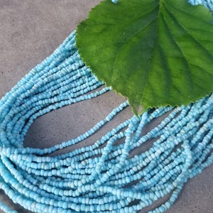 Könnte beinhalten: Eine Sammlung hellblauer Perlenketten. Ein großes grünes Blatt liegt auf den Halsketten. Die Perlen sind klein und zylindrisch, und die Halsketten sind in einem geschichteten Muster angeordnet.