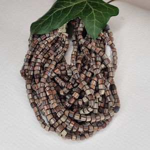 Könnte beinhalten: Ein Haufen brauner, weißer und orangefarbener quadratischer Perlen. Die Perlen sind in einem Kreis angeordnet, mit einem grünen Blatt obenauf.