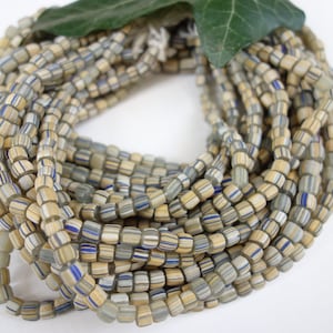 Könnte beinhalten: Eine Strähne kleiner, quadratischer Glasperlen. Die Perlen sind in einem Mix aus Beige, Weiß und Blau gehalten. Die Perlen sind kreisförmig angeordnet.