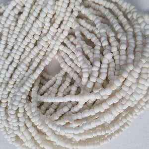 Könnte beinhalten: Nahaufnahme eines Haufens weißer, quadratisch geformter Perlen. Die Perlen sind aufgefädelt und bilden lange Stränge. Die Perlen haben eine leicht strukturierte Oberfläche und sind dicht gepackt, wodurch ein dichtes, undurchsichtiges Aussehen entsteht. Ideal für die Schmuckherstellung.