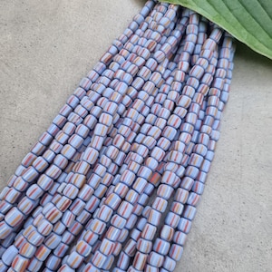 Op de afbeelding: Een streng lichtblauwe glazen kralen met witte en oranje strepen. De kralen zijn klein en cilindrisch van vorm.