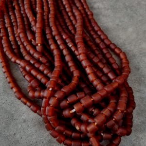 Könnte beinhalten: Eine Ansammlung mehrerer Stränge aus kleinen, zylindrischen Perlen mit mattem Finish in einem tiefen, erdigen Terrakotta-Rot. Die Perlen sind auf einen dünnen, hellen Faden aufgezogen und bilden ein dichtes, strukturiertes Bündel.