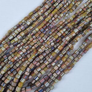 Può includere: Una fila di perline di vetro colorate con un motivo a strisce gialle, bianche, rosse, blu e marroni. Le perline sono quadrate e hanno una finitura lucida.