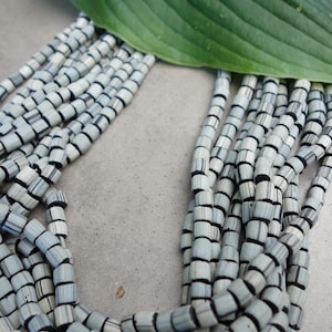 Könnte beinhalten: Eine mehrsträngige Halskette mit zylindrischen Perlen in Grau und Schwarz. Die Perlen sind in mehreren Strängen angeordnet, wodurch ein geschichteter Effekt entsteht. Ein großes grünes Blatt liegt oben auf der Halskette.