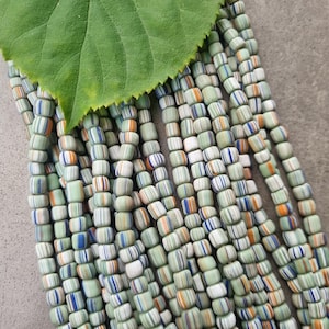 Könnte beinhalten: Stränge aus zylindrischen Perlen mit grünen, weißen, blauen und orangefarbenen Streifen. Ein großes grünes Blatt liegt auf den Perlen.