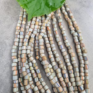 Könnte beinhalten: Nahaufnahme von mehreren Strängen zylindrischer Perlen. Die Perlen sind weiß mit orangefarbenen, blauen und beigefarbenen Streifen. Oben befindet sich ein grünes Blatt. Diese Perlen eignen sich für die Schmuckherstellung. Jede Perle ist etwa 0,5 cm lang.