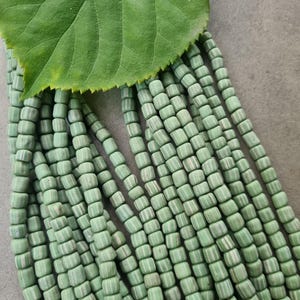 Op de afbeelding: Close-up van meerdere strengen lichtgroene, rechthoekige kralen met witte strepen. Een groot groen blad ligt bovenop de kralen. De kralen zijn waarschijnlijk voor het maken van sieraden. De kralen zijn ongeveer 0,5 cm lang.
