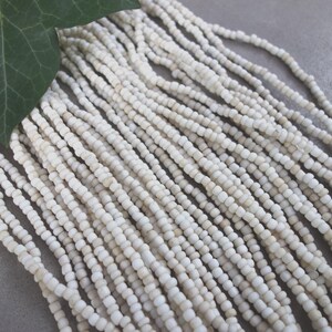 Könnte beinhalten: Stränge aus kleinen, runden, cremefarbenen Perlen. Oben links ist ein grünes Blatt zu sehen. Die Perlen scheinen aus einem natürlichen Material wie Stein oder Muschel zu bestehen.