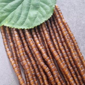 Könnte beinhalten: Nahaufnahme von Strängen aus bernsteinfarbenen, quadratisch geformten Perlen. Ein großes grünes Blatt bedeckt die Perlen teilweise. Die Perlen sind in parallelen Linien angeordnet und bilden eine strukturierte Oberfläche. Der Hintergrund ist ein neutrales Grau.