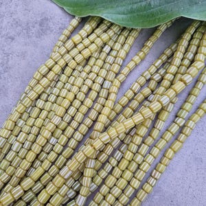 Könnte beinhalten: Eine Strähne gelber und weißer, gestreifter Glasperlen. Die Perlen sind zylindrisch und haben eine matte Oberfläche.