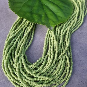 Könnte beinhalten: Eine mehrsträngige Halskette aus kleinen, hellgrünen Perlen. Ein großes, grünes Blatt liegt auf der Halskette. Die Perlen sind aufgefädelt und erzeugen einen drapierten Effekt. Die Halskette wird auf einer grauen Oberfläche präsentiert.