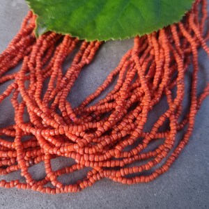Könnte beinhalten: Eine mehrsträngige Halskette aus zahlreichen kleinen, leuchtend orangefarbenen Saatperlen. Die Stränge sind in einer lockeren, fließenden Anordnung zusammengefasst und werden am oberen Rand teilweise von einem großen, gezackten grünen Blatt bedeckt.