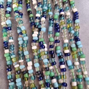 Peut inclure: Gros plan d'un collier multi-brins composé de perles bleues, vertes, blanches et transparentes. Les perles sont petites et de taille uniforme, créant un aspect texturé et coloré.