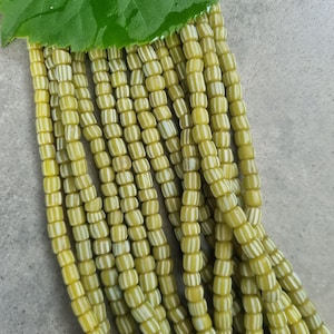 Könnte beinhalten: Stränge aus gelb-weiß gestreiften Perlen, mit einem grünen Blatt oben. Die Perlen sind zylindrisch und weisen ein sich wiederholendes Muster aus gelben und weißen Streifen auf. Diese Perlen werden wahrscheinlich zur Schmuckherstellung verwendet.