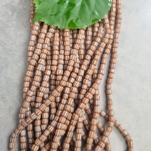 Könnte beinhalten: Stränge aus beige-weiß gestreiften Perlen, mit einem grünen Blatt obenauf. Die zylindrischen Perlen sind aufgefädelt. Die neutrale Farbe der Perlen macht sie für die Schmuckherstellung geeignet. Die Perlen haben einen Durchmesser von etwa 0,5 cm.