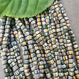 Könnte beinhalten: Mehrere Stränge bunter, gestreifter Perlen in Grün, Weiß, Gelb und Blau. Die Perlen sind zylindrisch und in mehreren Strängen angeordnet. Ein großes grünes Blatt liegt auf den Perlen.