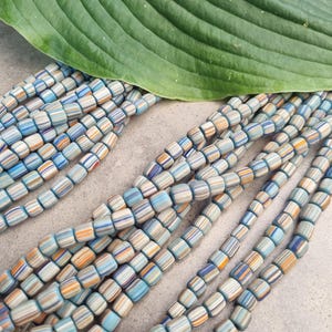 Op de afbeelding: Een streng gekleurde vierkante kralen met blauwe, oranje en witte strepen. De kralen zijn gemaakt van keramiek en hebben een glanzende afwerking.