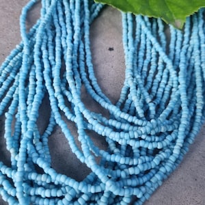 Könnte beinhalten: Nahaufnahme einer Halskette aus zahlreichen Strängen hellblauer Perlen. Ein grünes Blatt liegt auf den Perlen. Die Perlen sind klein und zylindrisch und erzeugen eine strukturierte Oberfläche. Die Halskette ist wahrscheinlich handgefertigt.