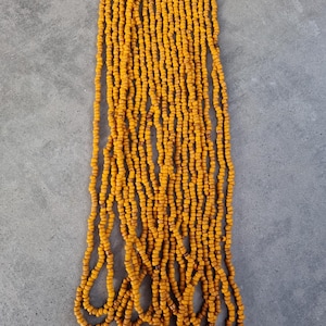 Op de afbeelding: Een lange streng oranje zaadkraaltjes. De kralen zijn klein en rond en zijn aan elkaar geregen op een dun draad. De streng ligt op een grijs oppervlak.