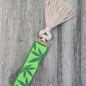 May include: Green bookmark with a marijuana leaf pattern and a beige tassel. The bookmark has a gold-colored metal top.