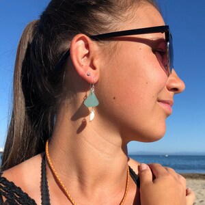 May include: A woman wearing a set of beach-themed jewelry. The set includes a necklace, earrings, and a bracelet, all featuring light blue sea glass pendants and silver accents. The jewelry is paired with a tan leather cord.
