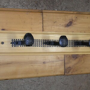 May include: A wooden coat rack with five black metal hooks shaped like railroad spikes. The hooks are mounted on a wooden board with a printed design of a railroad track.