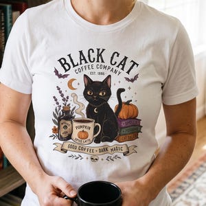 Könnte beinhalten: Ein weißes Kurzarm-T-Shirt mit einer Grafik eines schwarzen Katers, einer Tasse mit der Aufschrift Pumpkin Spice Latte, einer Zaubertrankflasche, einem Bücherstapel und Fledermäusen. Der Text lautet Black Cat Coffee Company Est 1886 und Good Coffee Dark Magic.
