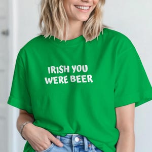 Könnte beinhalten: Ein leuchtend grünes T-Shirt mit dem weißen Text "IRISH YOU WERE BEER". Das Oberteil wird mit hellblauen Jeans kombiniert. Das Outfit hat eine entspannte, lässige Ästhetik, die für den Alltag geeignet ist.