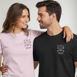Könnte beinhalten: Ein hellrosa und ein schwarzes T-Shirt mit einem grafischen Design auf der Brust. Das Design enthält Pfeile, Buchstaben, Zahlen und das Jahr 2021. Das Paar umarmt sich.
