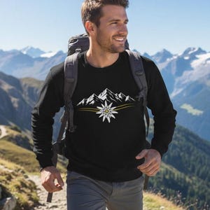 Könnte beinhalten: Schwarzes Langarmshirt mit weißem Berg- und Blumendesign. Die Person trägt das Shirt beim Wandern in den Bergen. Das Design beinhaltet einen gelben Streifen unter der Bergkette und der Blume.