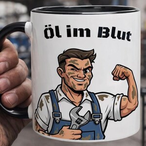 Puede incluir: Taza de cerámica blanca con asa y borde negros, con un mecánico de dibujos animados flexionando el brazo, sosteniendo una llave y cubierto de grasa. El texto "Öl im Blut" está impreso encima del mecánico.