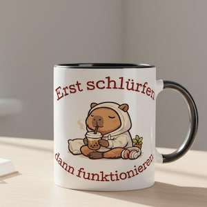 Può includere: Tazza in ceramica bianca con manico e bordo neri, con un capibara dei cartoni animati con cappuccio che beve da una tazza. La tazza ha il testo "Erst schlürfen dann funktionieren" in marrone. Un libro, occhiali e una piccola pianta sono sullo sfondo.