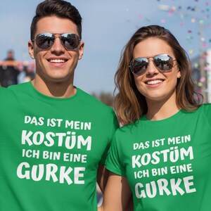 Puede incluir: Dos personas con camisetas verdes con el texto alemán "DAS IST MEIN KOSTÜM ICH BIN EINE GURKE" en blanco. El texto se traduce como "Este es mi disfraz, soy un pepinillo". Ambos llevan gafas de sol y saludan.