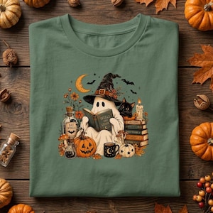 Könnte beinhalten: Ein salbeigrünes T-Shirt mit einer Grafik eines Geistes, der einen Hexenhut trägt und ein Buch mit dem Titel Spells and Stories liest. Die Illustration zeigt zudem eine schwarze Katze, eine Mondsichel, Kürbisse, eine Flasche und Bücher.