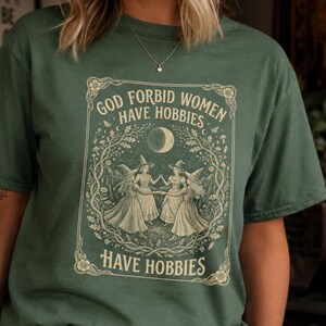 Op de afbeelding: Een bosgroen T-shirt met korte mouwen en een crèmekleurige grafische print van drie figuren met puntmutsen die in een cirkel dansen onder een maansikkel, omringd door bloemmotieven en de tekst God Forbid Women Have Hobbies.