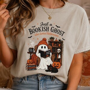 Könnte beinhalten: Ein hellbeiges T-Shirt mit einer Grafik eines Geistes mit orangefarbener Mütze, der in einem Sessel sitzt und ein Buch mit dem Titel Spooky Stories liest. Der Text lautet Just a BOOKISH GHOST who loves Halloween, ergänzt durch eine schwarze Katze, Kürbisse und ein Bücherregal.