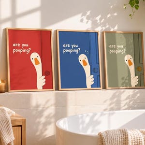 May include: Three framed wall art prints featuring a white goose peeking around a door with the text "are you pooping?" printed in white. The prints have red, blue, and sage green backgrounds, each displayed in a light-colored wooden frame.