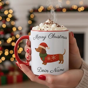 Op de afbeelding: Een witte keramische mok met een rood handvat, voorzien van een afbeelding van een teckel met een kerstmuts, een groene sjaal en een rode trui met witte sneeuwvlokken, en de tekst Merry Christmas en Dein Name, gevuld met marshmallows en cacao.