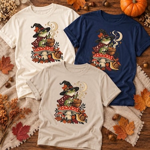 Könnte beinhalten: Drei kurzärmelige T-Shirts in Creme, Marineblau und Hellbeige, jedes mit einer Grafik eines grünen Frosches, der einen Hexenhut und Umhang trägt, auf einem roten Pilz sitzt, in einem Topf rührt und von einer Schnecke sowie Herbstblättern begleitet wird.