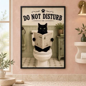 Könnte beinhalten: Ein gerahmter Kunstdruck, der eine schwarze Katze zeigt, die auf einer weißen Toilette sitzt und eine Zeitung liest. Der Text DO NOT DISTURB ist oben in Schwarz über einem Pfotenabdruck-Symbol gedruckt. Der Hintergrund zeigt ein Badezimmer mit hellgrüner Wandvertäfelung.
