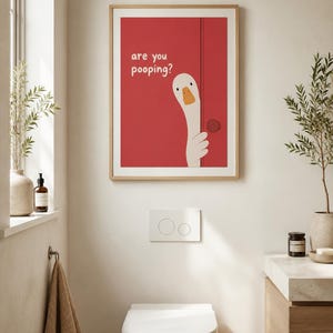 May include: A framed art print with a red background and a cartoon goose peeking out from behind a door. The text "are you pooping?" is above the goose. The print hangs above a white toilet in a bathroom.