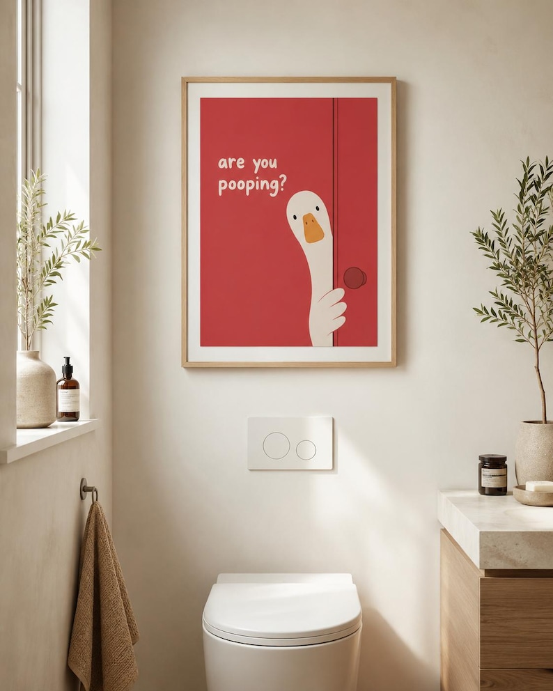 May include: A framed art print with a red background and a cartoon goose peeking out from behind a door. The text "are you pooping?" is above the goose. The print hangs above a white toilet in a bathroom.
