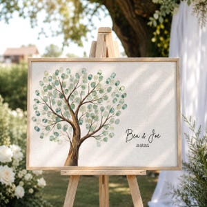 Op de afbeelding: Een rechthoekige canvasprint op een houten ezel met een bruine boomstam en talloze groene en blauwgroene vingerafdruk-achtige bladeren. De tekst "Bea & Joe" en de datum "20.08.2026" zijn in zwarte inkt aan de rechterkant van het canvas gedrukt.