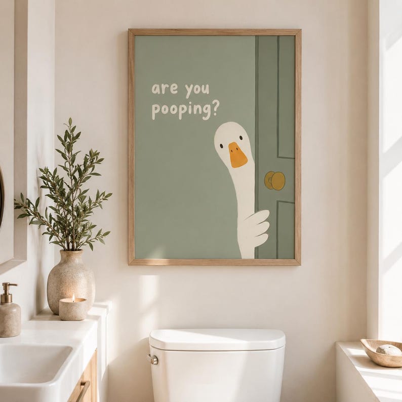 May include: A framed print featuring a cartoon goose peeking from behind a green door. The text "are you pooping?" is above the goose. The print has a light wood frame and is displayed in a bathroom setting.