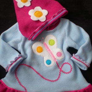 Puede incluir: Un vestido de capucha de forro polar azul claro con un ribete de volantes rosa. El vestido presenta una aplicación de mariposa con detalles coloridos y un ribete de cuentas rosa y azul. La capucha es rosa con flores de fieltro blanco.