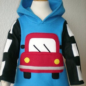 Op de afbeelding: Een blauwe fleece hoodie met zwarte en witte mouwen en een rode auto-applicatie op de voorkant. De auto heeft gele koplampen en een witte voorruit.