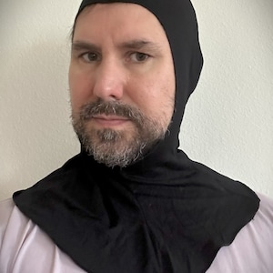 Puede incluir: Una capucha negra con cubrecuello. La capucha está hecha de una tela suave y oscura y se usa alrededor de la cabeza y el cuello. La capucha es de color negro liso.